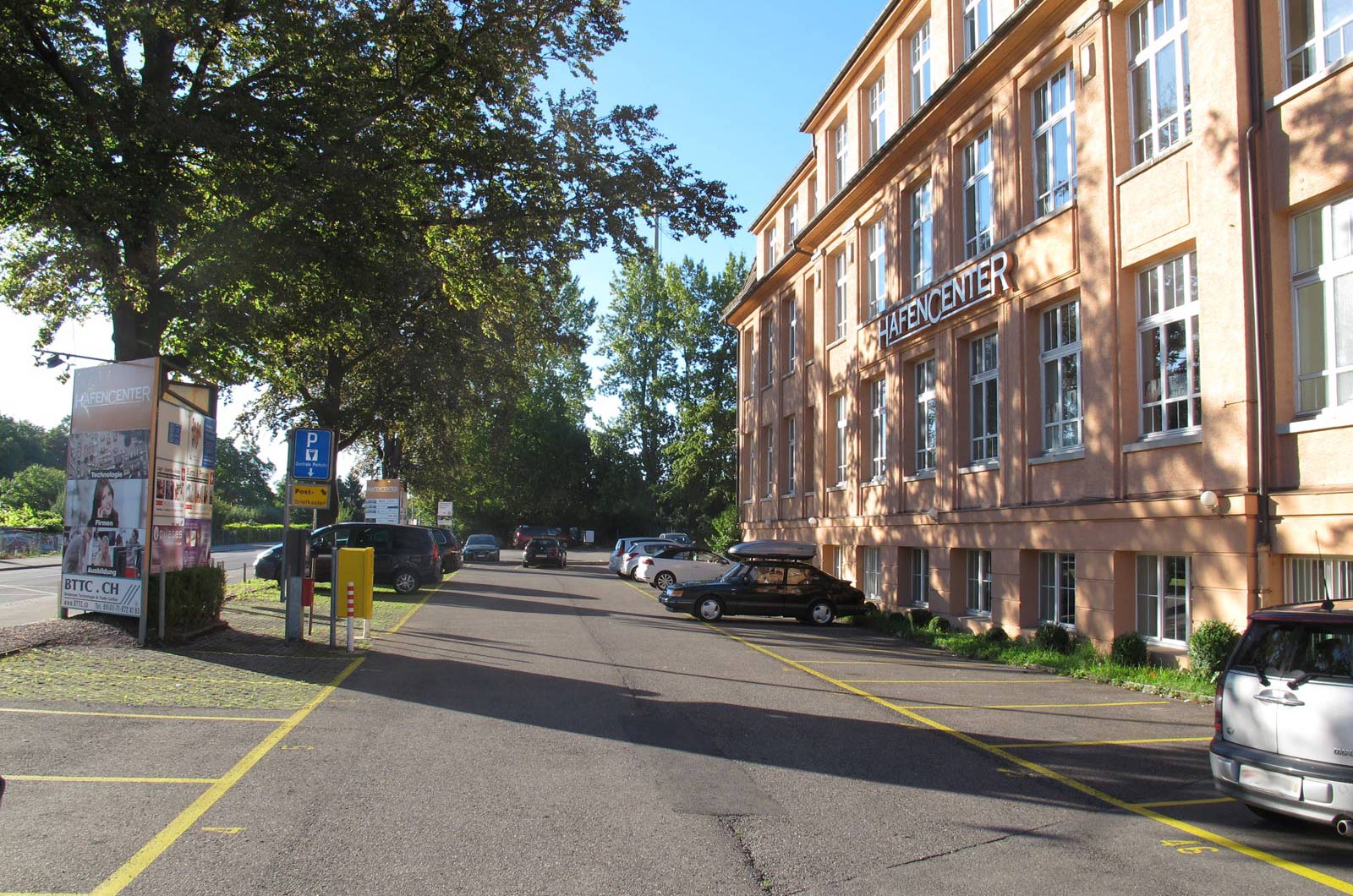 Parkplatzbereich vor dem Gebäude Hafencenter mit mehreren geparkten Fahrzeugen und Bäumen.